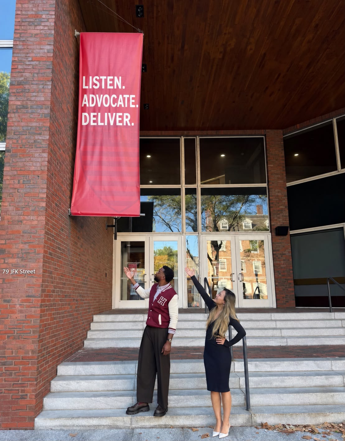 Xinyi and Andrew on the steps of 79 JFK Street, reaching toward a crimson banner reading “Listen. Advocate. Deliver.”