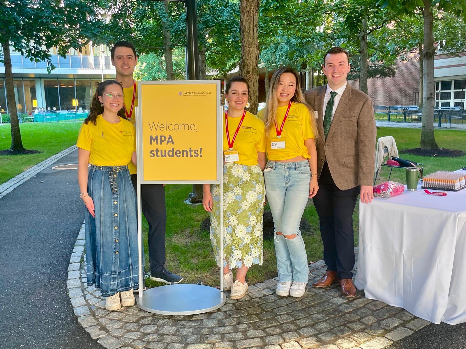 Orientation Leaders in yellow HKS tees beside a "Welcome, MPA students!" sign